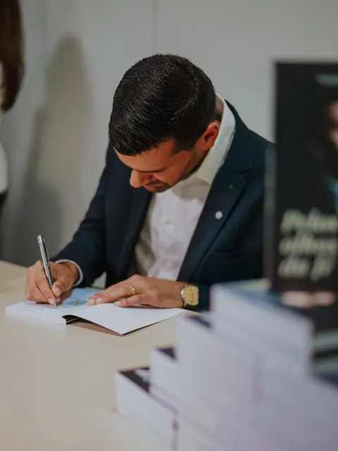 Serginho Vidigal autografando um exemplar do livro Pelos Olhos da Fé, com pilhas do livro à frente